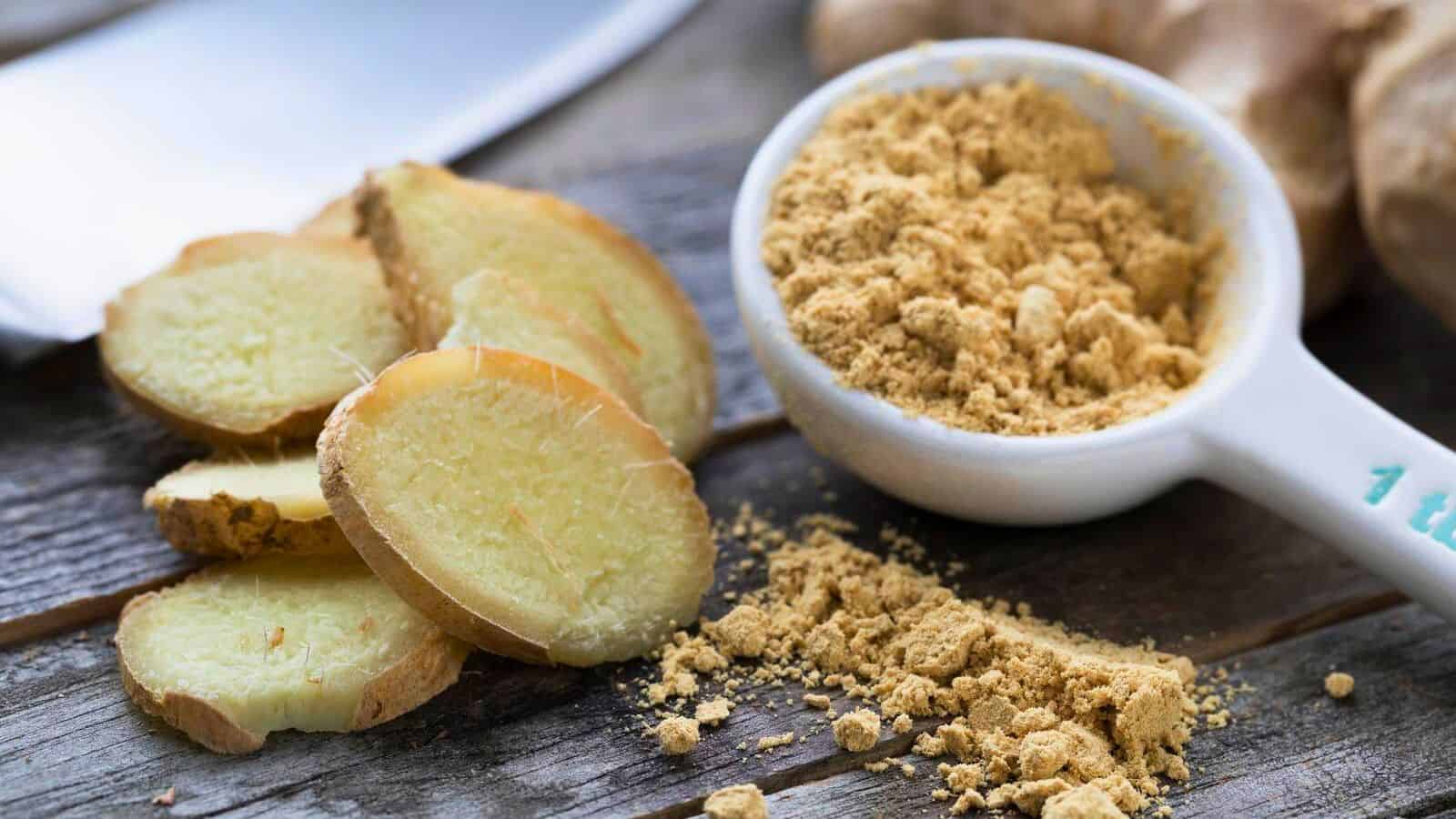 A close-up image shows several slices of fresh ginger root, a small heap of ground ginger powder, and a metal measuring spoon filled with more ginger powder. Perfect spices for coffee, the background is a rustic wooden surface with a knife partially visible at the top left corner.