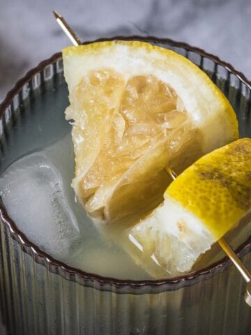 A close-up of a dark glass containing a drink with ice cubes and a wedge of lemon inside. A lemon wedge is also speared on a cocktail pick placed on the glass rim. Another glass with a lemon wedge is partially visible in the background.