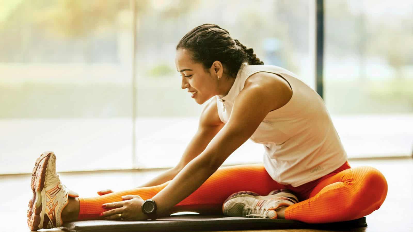 A person with braided hair, wearing an orange and white athletic outfit, is sitting on a mat indoors, stretching one leg forward while reaching for their foot with their hand. The background shows large windows with a view of a park. They're dedicated to fitness and have chosen to stop drinking alcohol for better health.