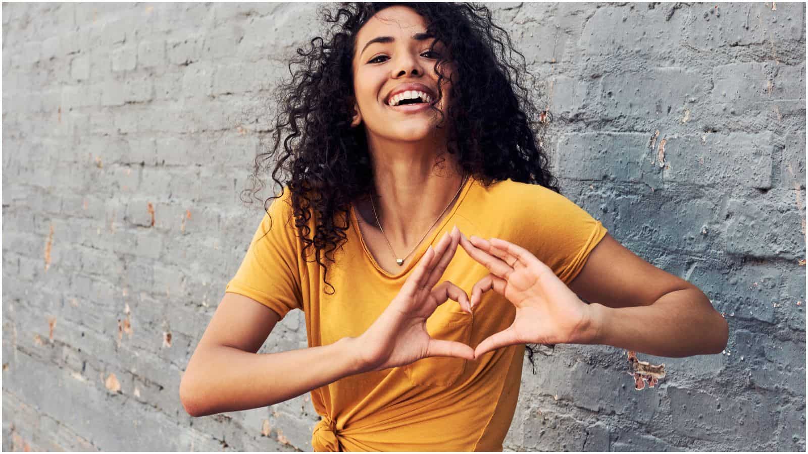 A person with curly hair, wearing a yellow shirt, stands in front of a gray brick wall. They are smiling and forming a heart shape with their hands, promoting the message to stop drinking alcohol.