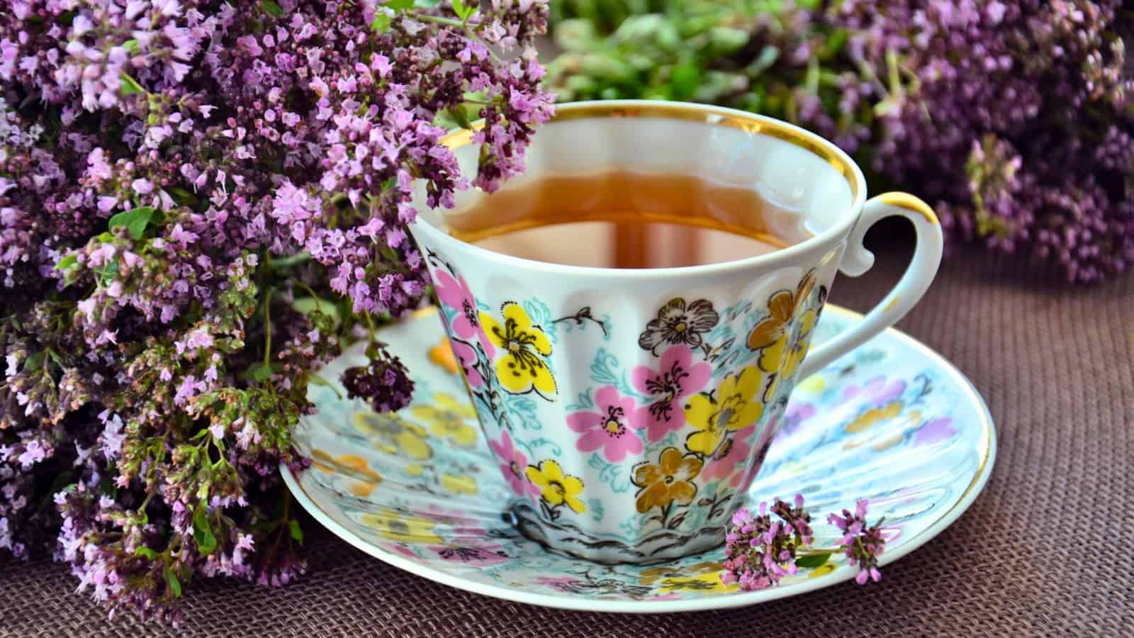 A floral-patterned teacup filled with tea sits on a matching saucer. The cup and saucer feature colorful flower designs. Surrounding the cup are sprigs of purple flowers on a brown fabric surface.