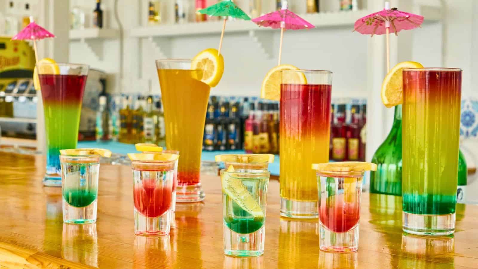 A bar counter displays various vibrant drinks, most with gradient colors from green to red and topped with lemon slices and umbrellas. In the background, there are bottles lined up on shelves, a subtle reminder of the types of alcohol you shouldn't be drinking. The setting has a casual and colorful ambiance.