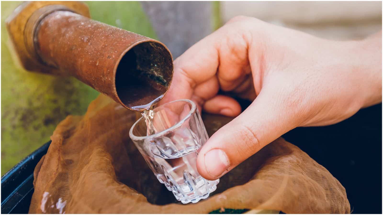 A hand holds a small glass underneath a rusty spout, collecting a clear liquid. The liquid flows steadily from the spout into the glass, hinting at an outdoor setup. This scene evokes caution, as it reminds one of the types of alcohol you shouldn't be drinking due to potential contaminants.