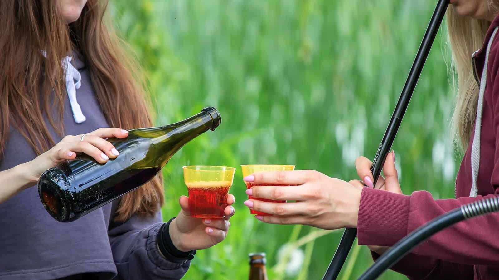 Two people wearing hoodies are holding plastic cups while one pours liquid from a beer bottle into them. The background is blurred greenery, and there are other beverage bottles on the table in front of them, highlighting some types of alcohol you shouldn't be drinking.