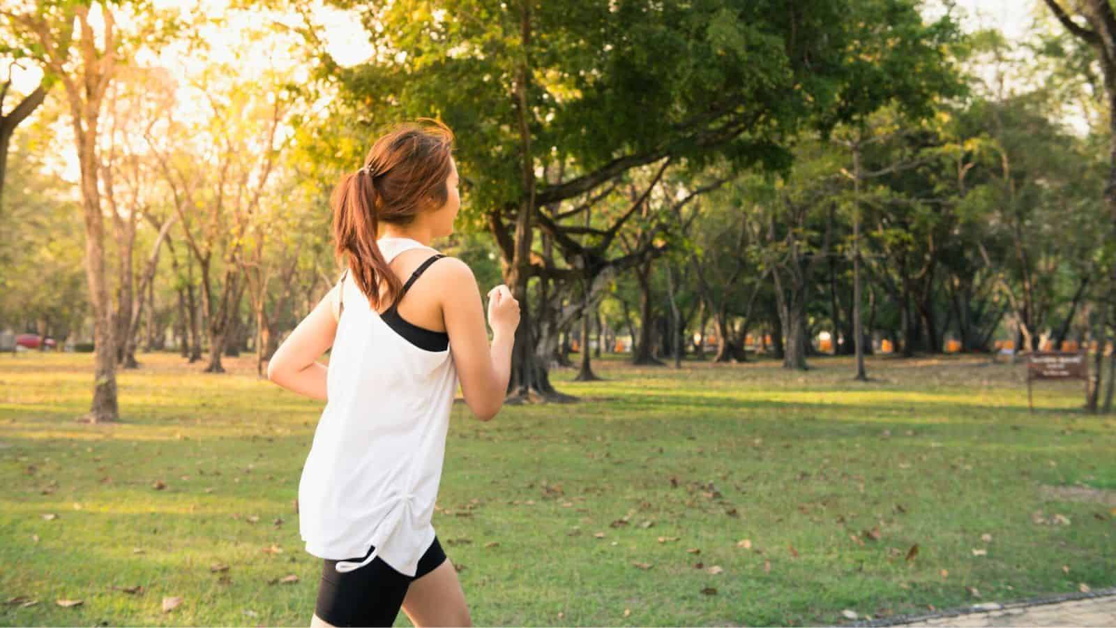 A person in a white sleeveless shirt and black shorts jogs on a path in a park with green grass and trees. The sun is either rising or setting, casting a warm light over the scene, reminiscent of the calming effect of lemon water before bed.