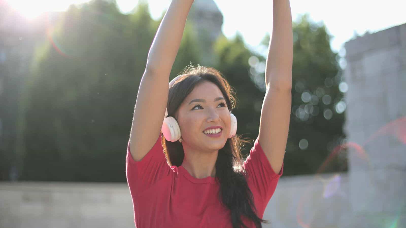 A person wearing pink headphones and a red shirt raises their arms while standing outdoors. The sun shines brightly in the background, creating a lens flare effect. Trees and a concrete structure are visible behind the person, reminding us how vibrant life can be when you stop drinking alcohol.