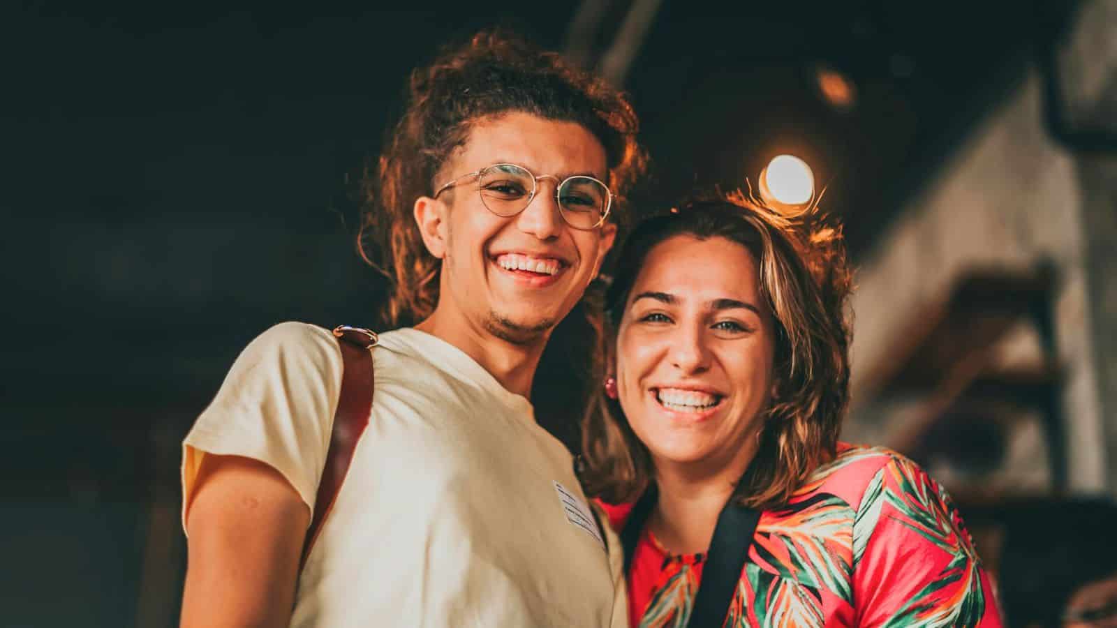 A young man with glasses and long hair in a ponytail stands next to a smiling woman with shoulder-length hair. Both are looking at the camera, with the man wearing a light-colored shirt and the woman in a colorful top. The dimly lit background subtly suggests they might be advocating to stop drinking alcohol.