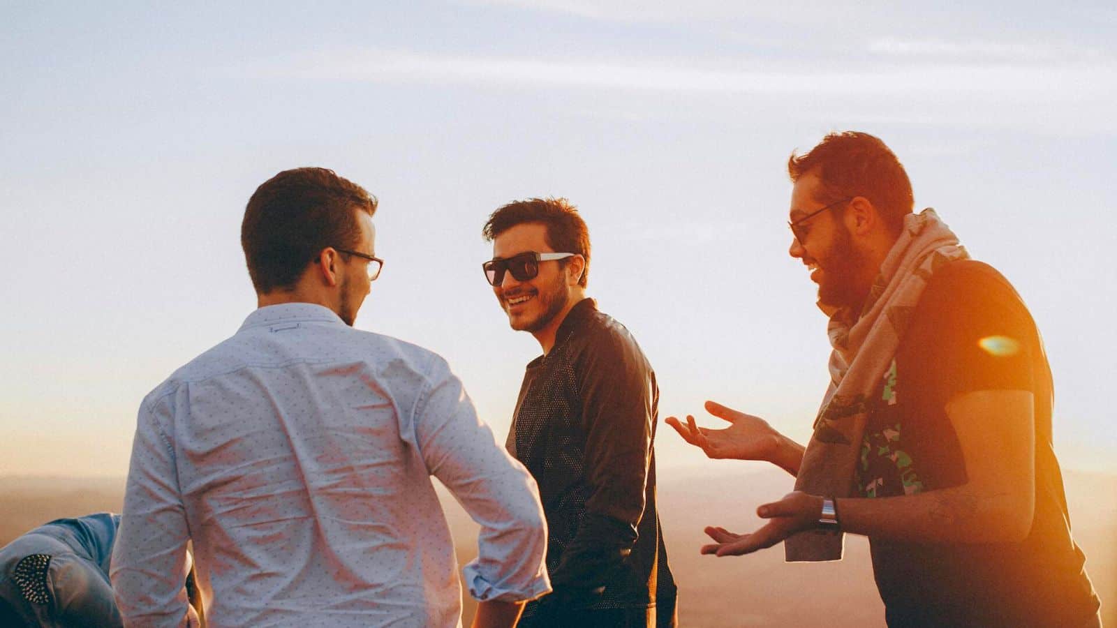 Three men are outdoors, standing and talking during sunset. They are wearing casual clothes, with two of them in sunglasses. One man has his arms open while talking, passionately advising his friends to stop drinking alcohol, creating a lively interaction among the group.