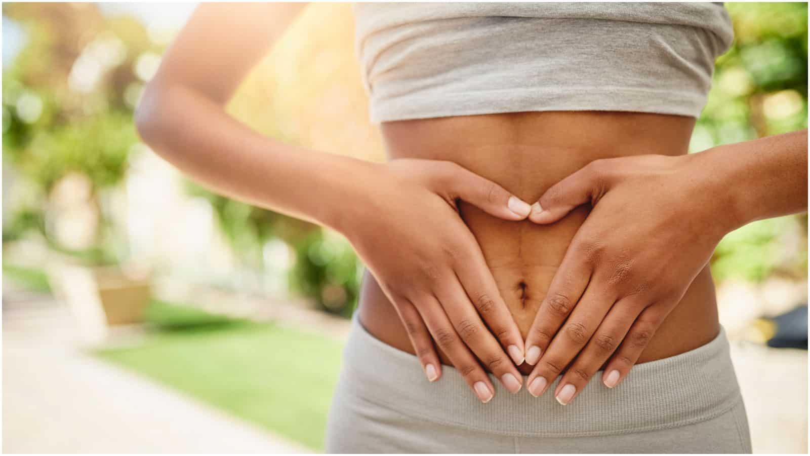 A person wearing a grey crop top and grey pants forms a heart shape with their hands over their bare midriff. The background is blurred with greenery and bright light.