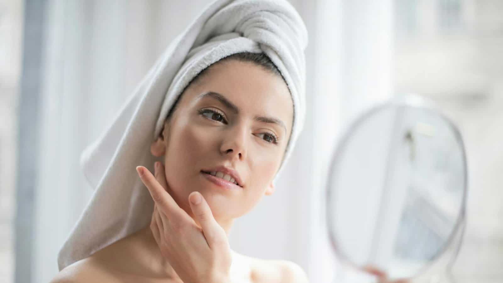 A person with a towel wrapped around their head is looking into a handheld mirror while gently touching their face. They are in a well-lit room with white curtains in the background.