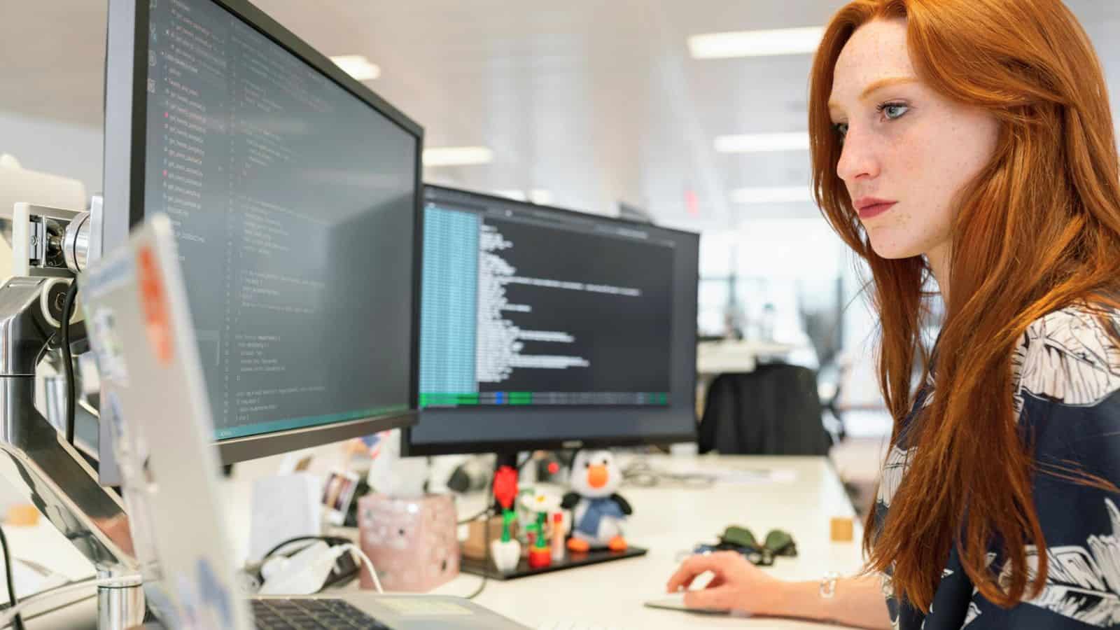 A woman with long red hair is working at a desk with two large computer monitors displaying coding and data. Amidst the office supplies and small figurines, a note saying "Stop Drinking Alcohol" serves as a motivational reminder. She is focused, sitting in an office environment.