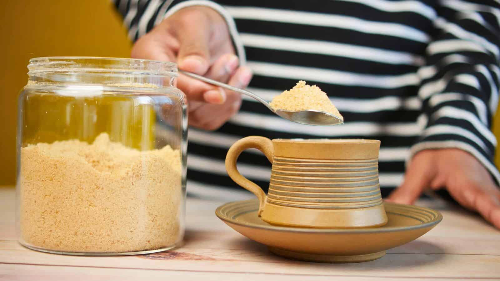 A person wearing a black and white striped shirt is seen holding a spoonful of brown sugar over a ceramic cup and saucer. To the left of the cup, there's a glass jar filled with brown sugar on a light wood surface&mdash;everything you need to know about simple syrup starts here.