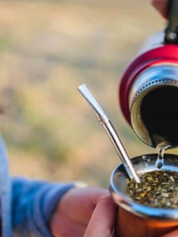 A person is pouring hot water from a thermos into a gourd filled with yerba mate, with a metal straw (bombilla) inside. Another person holding the gourd is partially visible. The background is blurred, highlighting an outdoor setting.