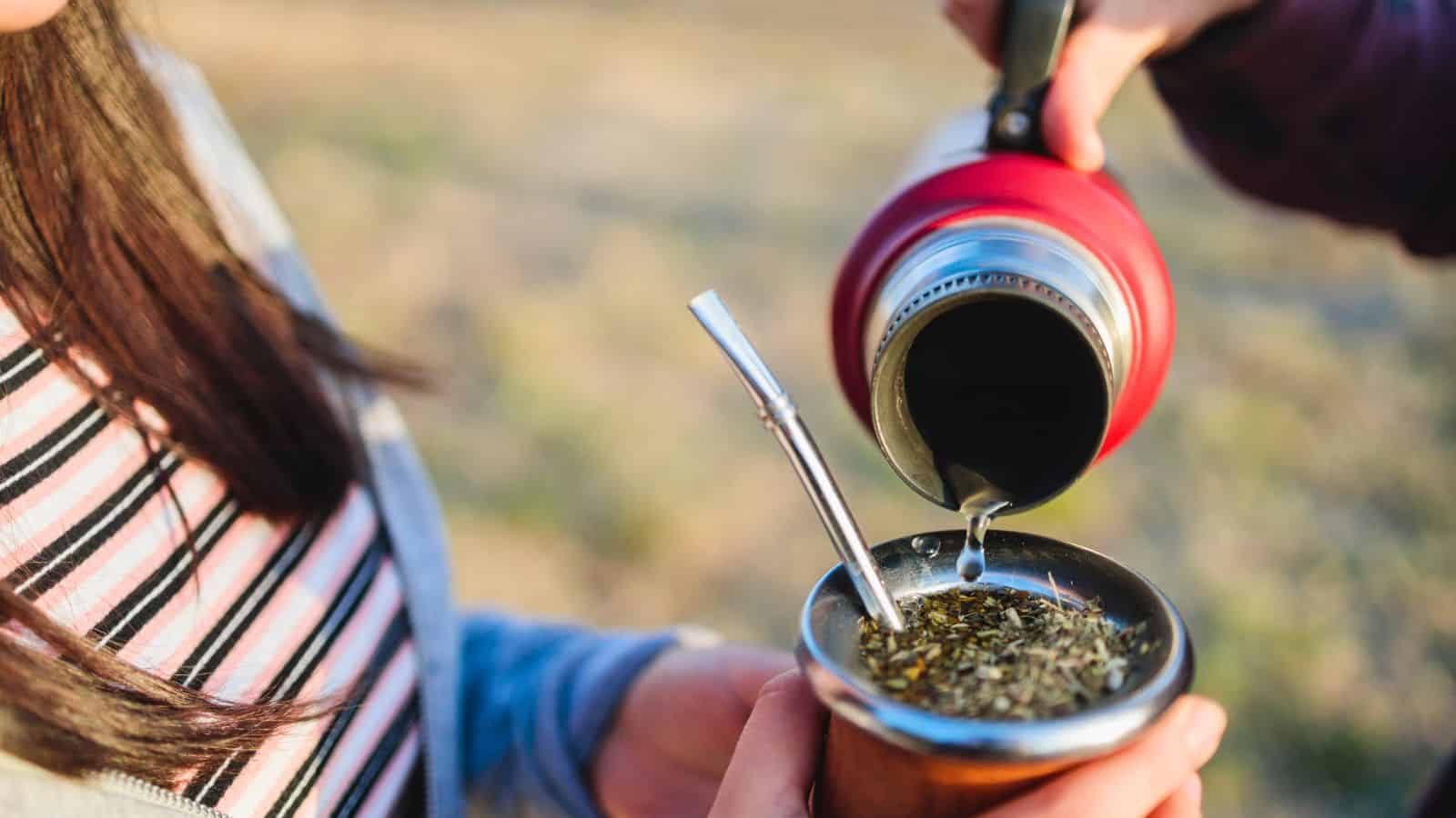 A person is pouring hot water from a thermos into a gourd filled with yerba mate, with a metal straw (bombilla) inside. Another person holding the gourd is partially visible. The background is blurred, highlighting an outdoor setting.