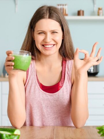 Woman doing the OK gesture while holding a glass of green smoothie on the other hand