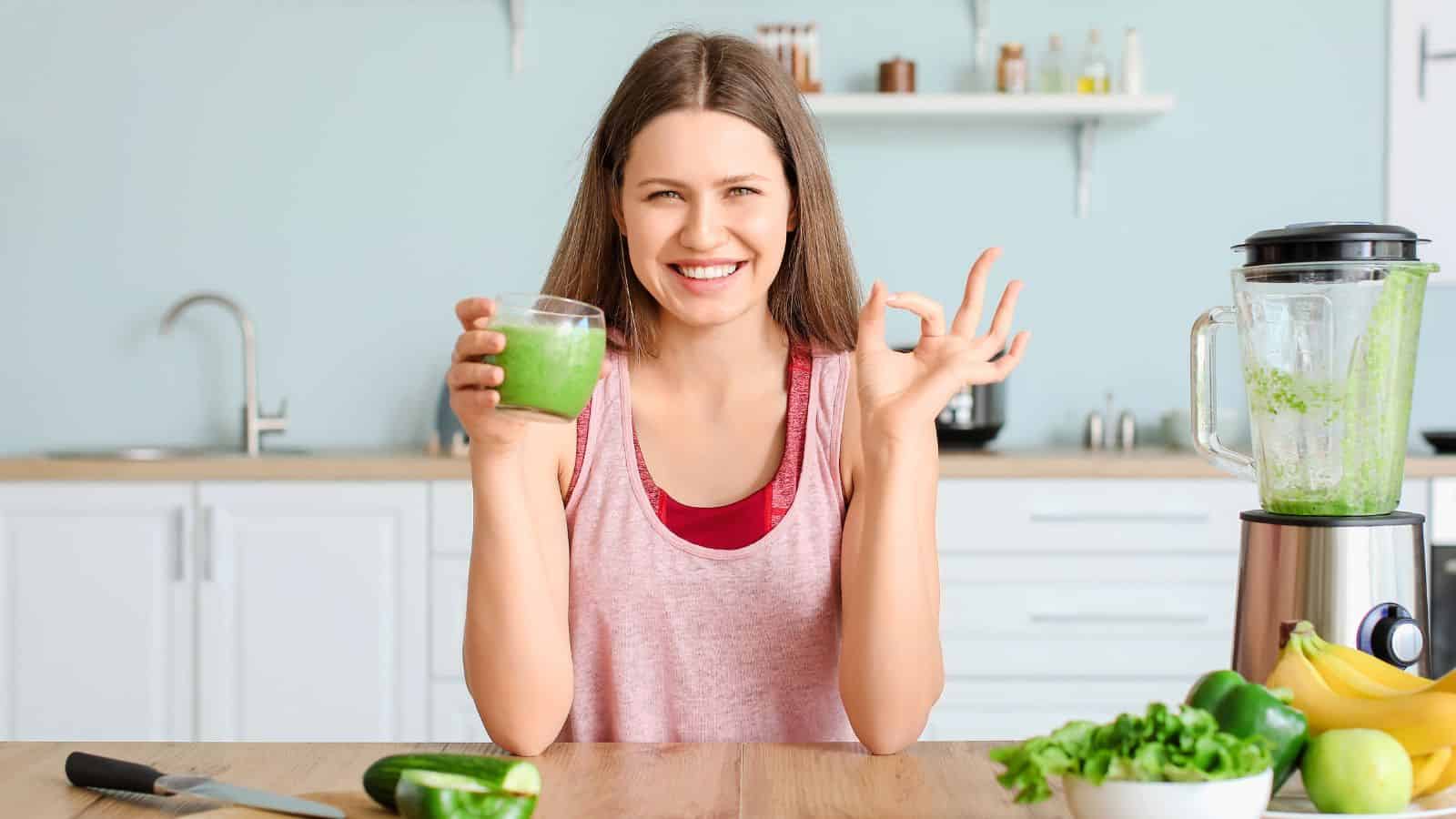 Woman doing the OK gesture while holding a glass of green smoothie on the other hand