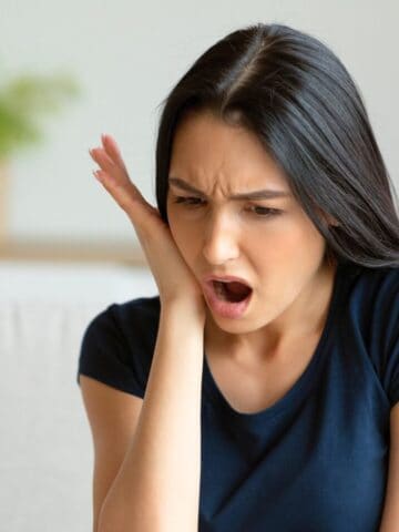 A woman with long black hair, wearing a dark blue shirt, is sitting and holding her face with one hand. She appears to be in pain or discomfort, perhaps from consuming alcoholic drinks that are bad for your teeth. Her mouth is open and eyebrows furrowed as a plant is visible in the blurred background.