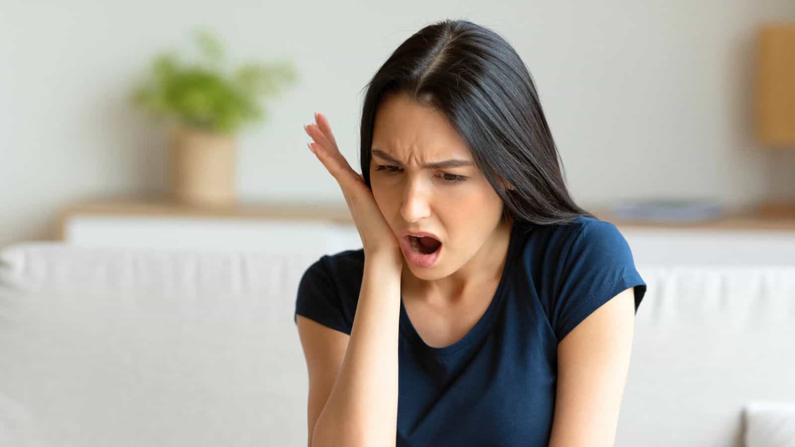 A woman with long black hair, wearing a dark blue shirt, is sitting and holding her face with one hand. She appears to be in pain or discomfort, perhaps from consuming alcoholic drinks that are bad for your teeth. Her mouth is open and eyebrows furrowed as a plant is visible in the blurred background.