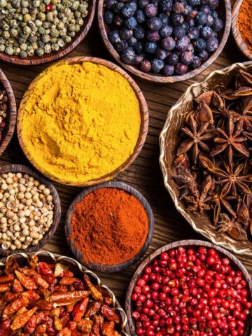 An assortment of various spices, including popular spices for coffee, in wooden bowls is displayed atop a wooden surface. The spices include turmeric, star anise, dried chili peppers, black peppercorns, paprika, coriander seeds, cloves, and more—showcasing a vibrant range of colors and textures.