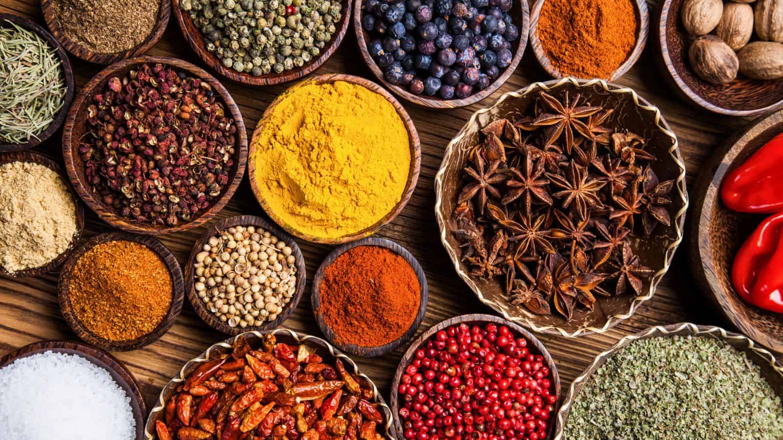 An assortment of various spices, including popular spices for coffee, in wooden bowls is displayed atop a wooden surface. The spices include turmeric, star anise, dried chili peppers, black peppercorns, paprika, coriander seeds, cloves, and more&mdash;showcasing a vibrant range of colors and textures.