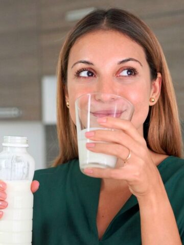 A woman with long brown hair is holding a glass of milk in one hand and a bottle of milk in the other. She is wearing a green shirt and standing in a kitchen with wooden cabinets in the background. She appears to be drinking or about to drink the milk, which are considered good drinks for gout.