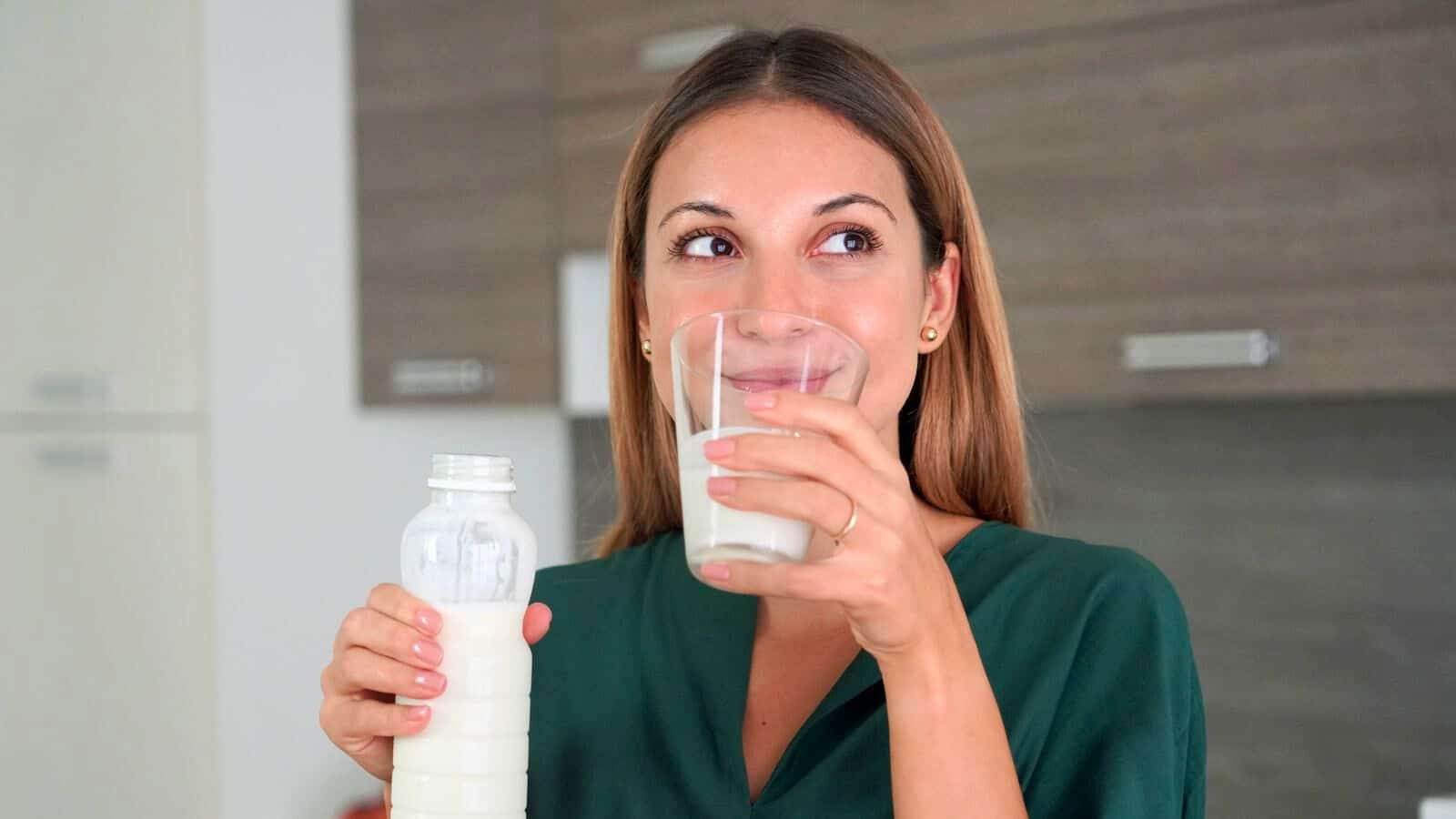 A woman with long brown hair is holding a glass of milk in one hand and a bottle of milk in the other. She is wearing a green shirt and standing in a kitchen with wooden cabinets in the background. She appears to be drinking or about to drink the milk, which are considered good drinks for gout.