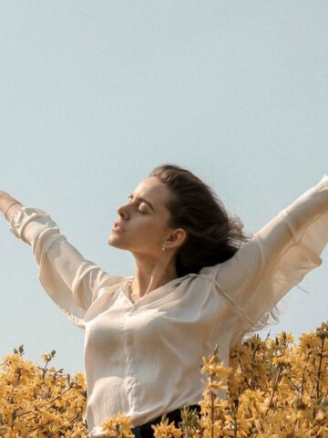 A person with long hair is standing in a field of yellow flowers with their arms raised and eyes closed, as if embracing a new chapter. They are wearing a white long-sleeved shirt. The sky above is clear and blue, symbolizing the clarity that comes when you stop drinking alcohol.