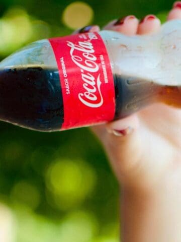 A person is sipping from a small, classic glass bottle of Coca-Cola. The bottle sports the iconic red label with white text. The background is outdoors, with green blurred foliage hinting at a sunny day. Among the list of sugary drinks, Coca-Cola stands out timelessly in the shot as their face remains mostly out of frame.