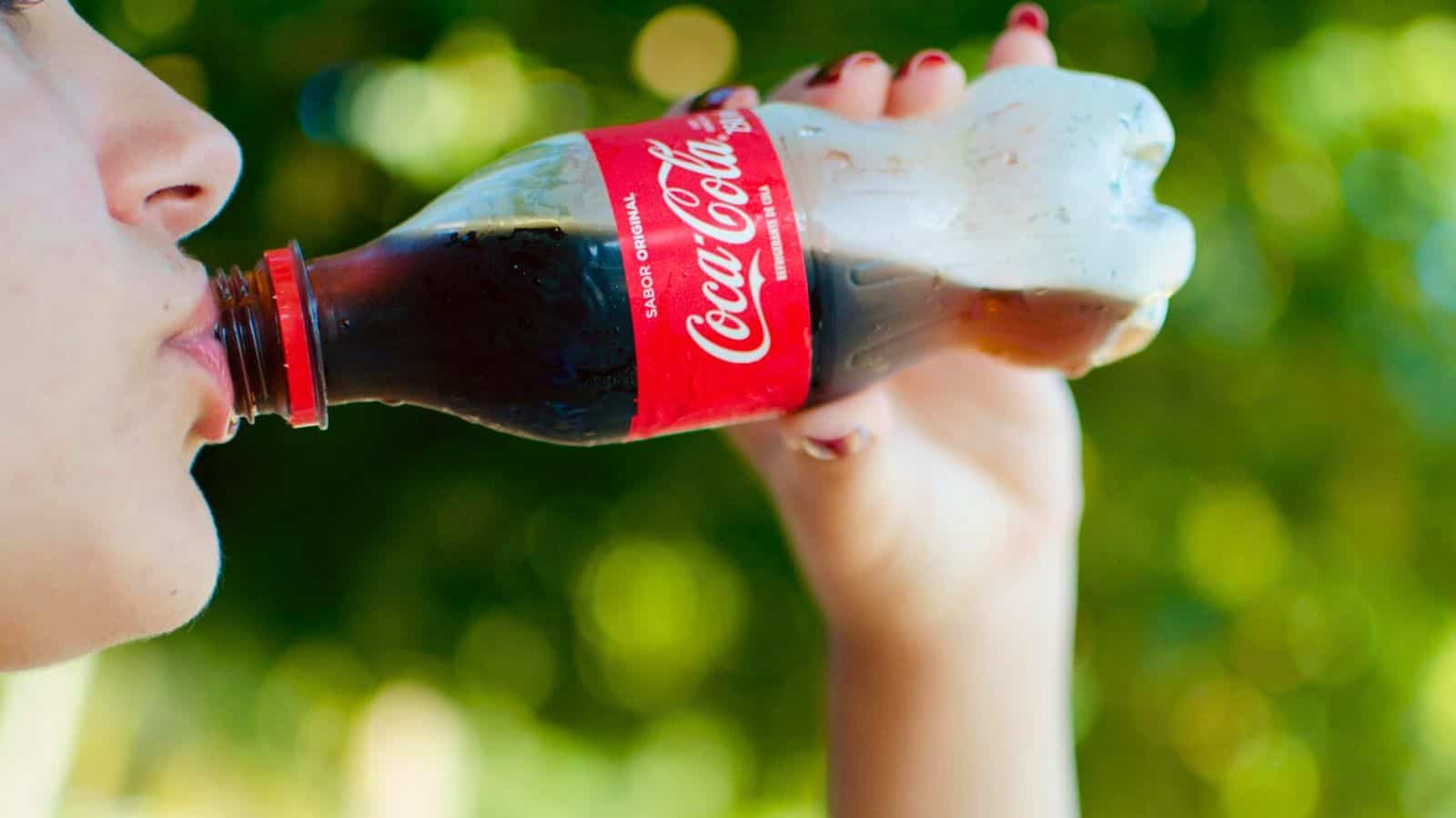 A person is sipping from a small, classic glass bottle of Coca-Cola. The bottle sports the iconic red label with white text. The background is outdoors, with green blurred foliage hinting at a sunny day. Among the list of sugary drinks, Coca-Cola stands out timelessly in the shot as their face remains mostly out of frame.