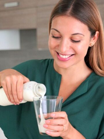 A woman with long brown hair is standing in a modern kitchen and smiling while pouring kefir from a glass bottle into a clear glass. Wearing a green blouse, she enjoys the benefits of kefir. Wooden cabinets and a white countertop are visible in the background.
