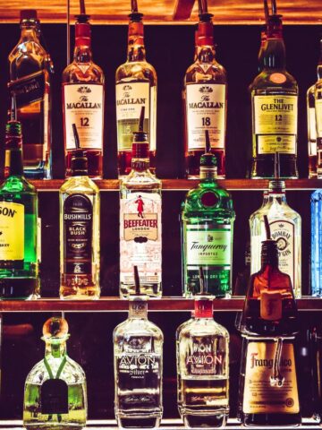 A bar shelf stocked with various types and brands of alcohol bottles, including whiskey, vodka, gin, tequila, rum, and others you should be cautious about. The bottles are arranged in multiple rows with varying shapes, sizes, and label colors.