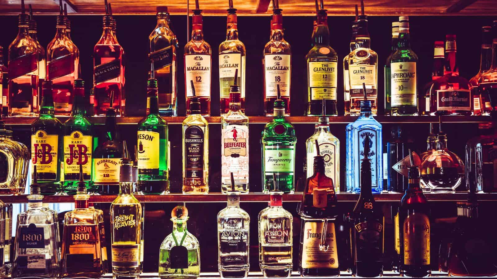 A bar shelf stocked with various types and brands of alcohol bottles, including whiskey, vodka, gin, tequila, rum, and others you should be cautious about. The bottles are arranged in multiple rows with varying shapes, sizes, and label colors.