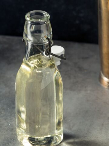 A clear glass bottle filled with a light-colored liquid sits on a gray countertop, offering everything you need to know about simple syrup. Next to it are a steel jigger and a tall metal shaker. Some salt or powder is scattered on the countertop. The background is dark and slightly out of focus.