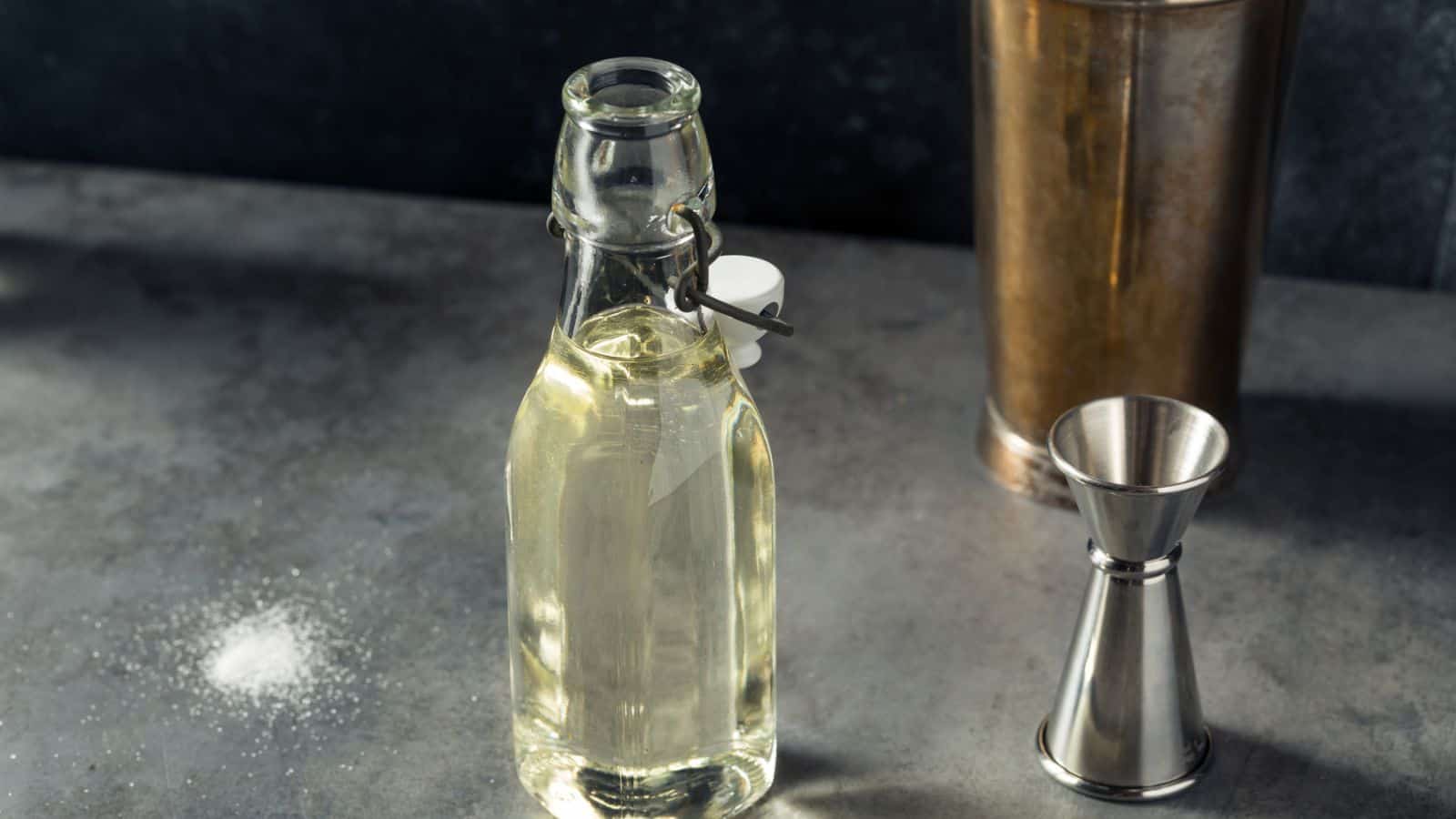 A clear glass bottle filled with a light-colored liquid sits on a gray countertop, offering everything you need to know about simple syrup. Next to it are a steel jigger and a tall metal shaker. Some salt or powder is scattered on the countertop. The background is dark and slightly out of focus.