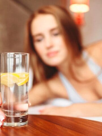 A woman with long hair, wearing a light top, is lying on a bed and reaching out to a glass of lemon water before bed. The glass is placed on a wooden bedside table. The background shows part of the bed and a lamp.