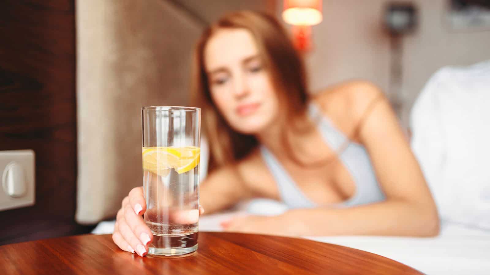 A woman with long hair, wearing a light top, is lying on a bed and reaching out to a glass of lemon water before bed. The glass is placed on a wooden bedside table. The background shows part of the bed and a lamp.