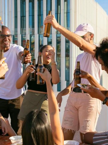 A group of people are on a rooftop, raising bottles in a celebratory toast. They are smiling and seem to be enjoying themselves while following alcohol rules. In the background, there is a modern building with vertical lines, and a view of the water.