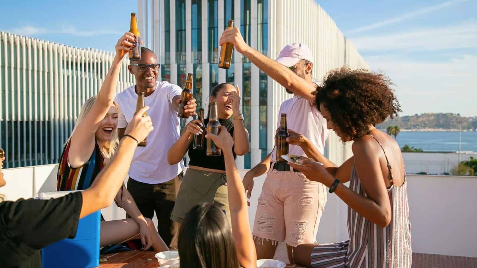 A group of people are on a rooftop, raising bottles in a celebratory toast. They are smiling and seem to be enjoying themselves while following alcohol rules. In the background, there is a modern building with vertical lines, and a view of the water.