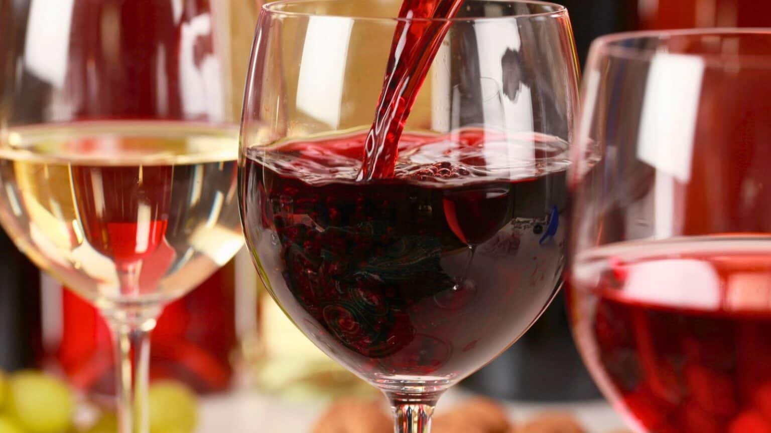 A close-up of three wine glasses: one with white wine, one being filled with a red wine varietal, and one with rosé. The background includes blurred colors and wine bottles. Grapes and corks are artfully arranged in the foreground.