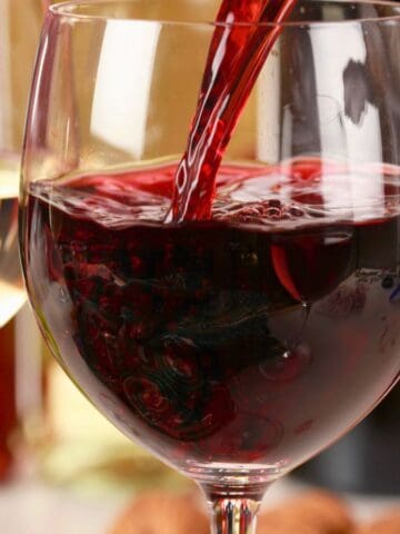 A close-up of three wine glasses: one with white wine, one being filled with a red wine varietal, and one with rosé. The background includes blurred colors and wine bottles. Grapes and corks are artfully arranged in the foreground.