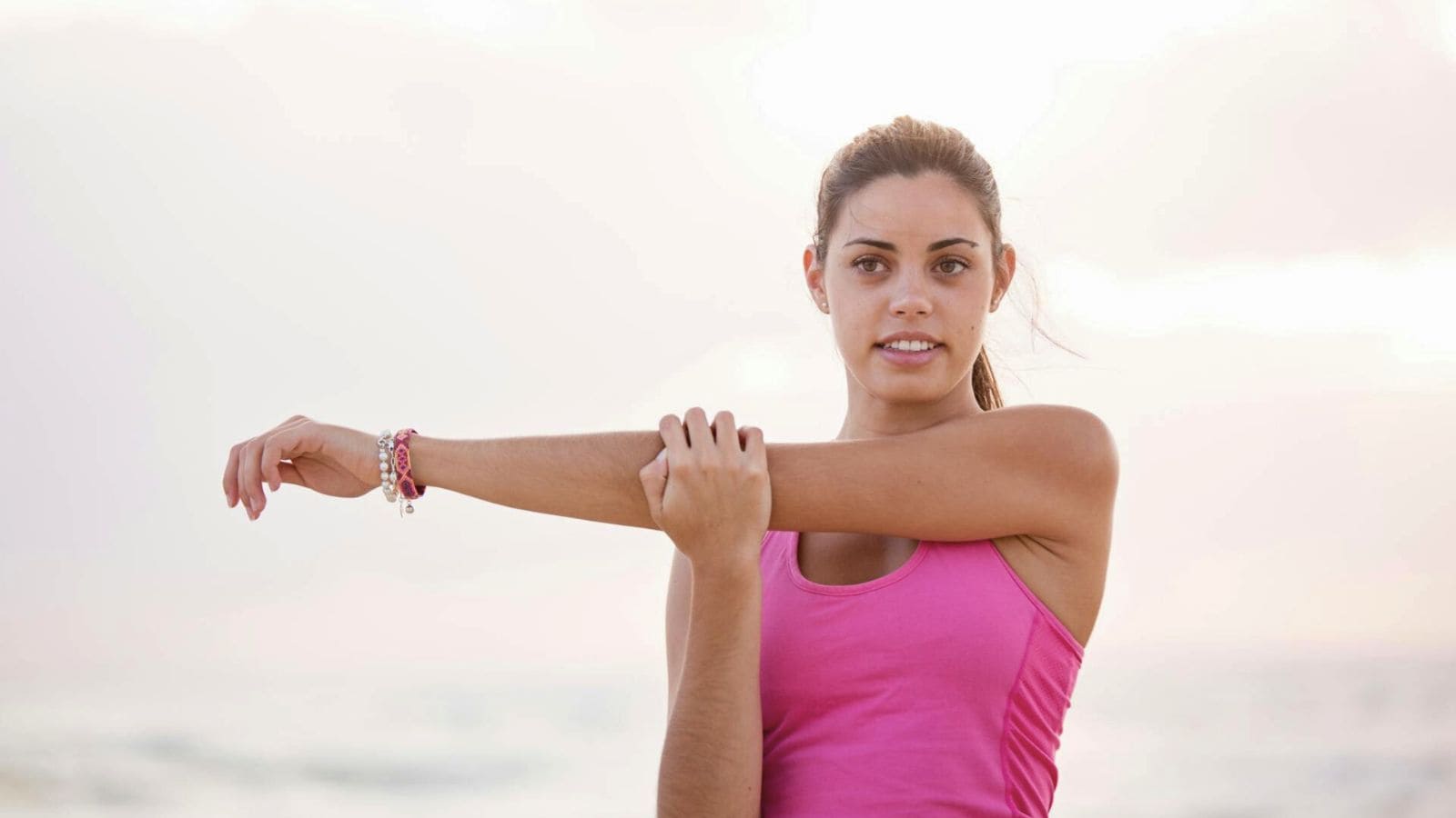 A person wearing a bright pink tank top stretches their left arm across their chest with their right hand, enjoying the benefits of lemon water before bed. They are outdoors, possibly at a beach, with a blurred background of the sea and sky. Their gaze is directed toward the camera.
