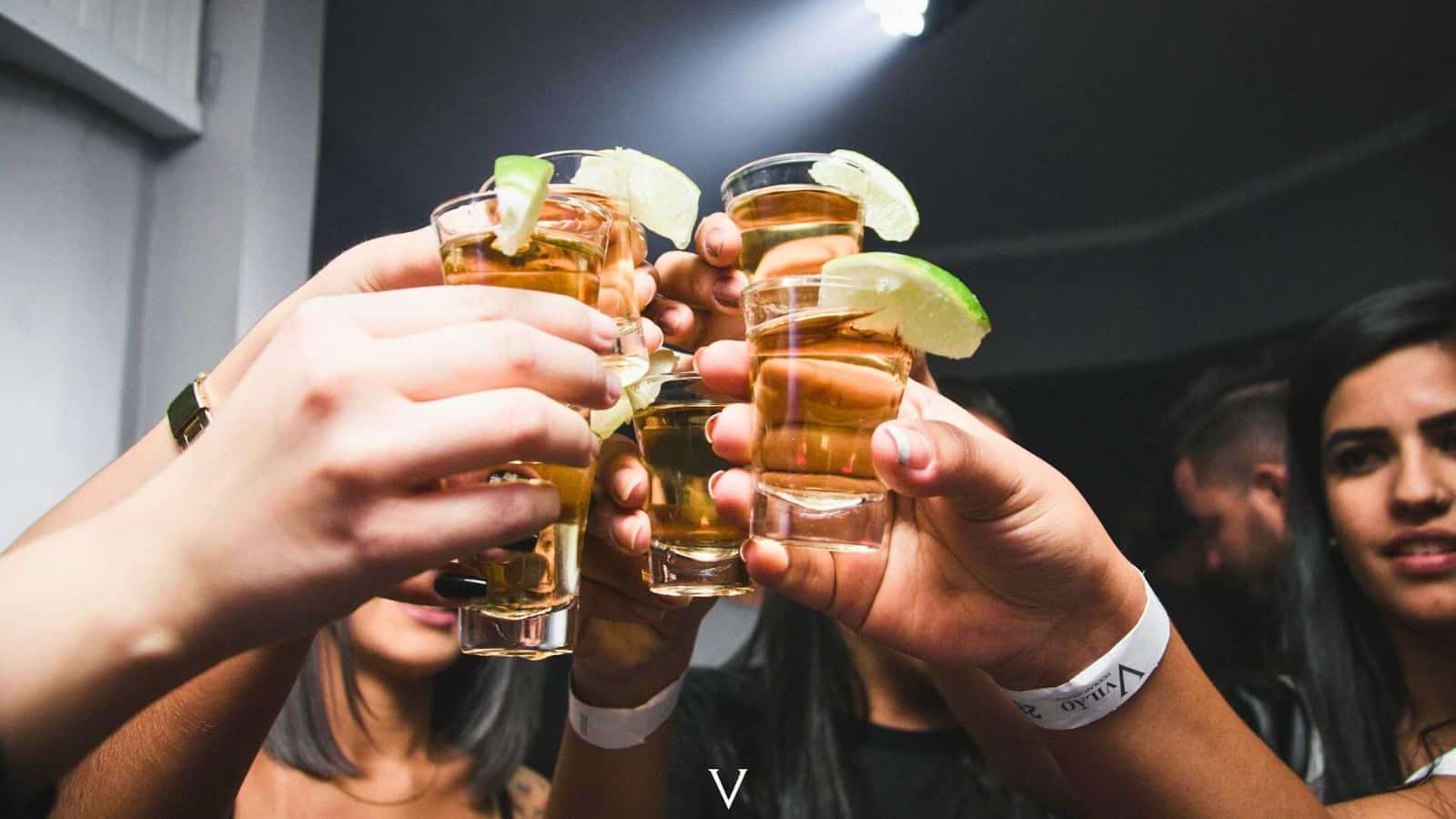 A group of people holding up shot glasses filled with a brown liquid, garnished with lime wedges. The background is dimly lit, and the focus is on the hands and shot glasses, all raised in celebration while adhering to alcohol rules.