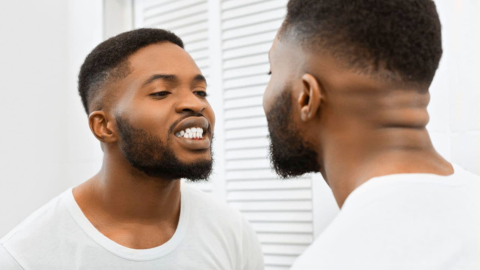 A man with short hair and a beard is looking at his reflection in the mirror. He is wearing a white shirt and appears to be examining his teeth, which are bared, perhaps checking for any damage caused by alcoholic drinks that are bad for your teeth. The background shows a white shuttered door.