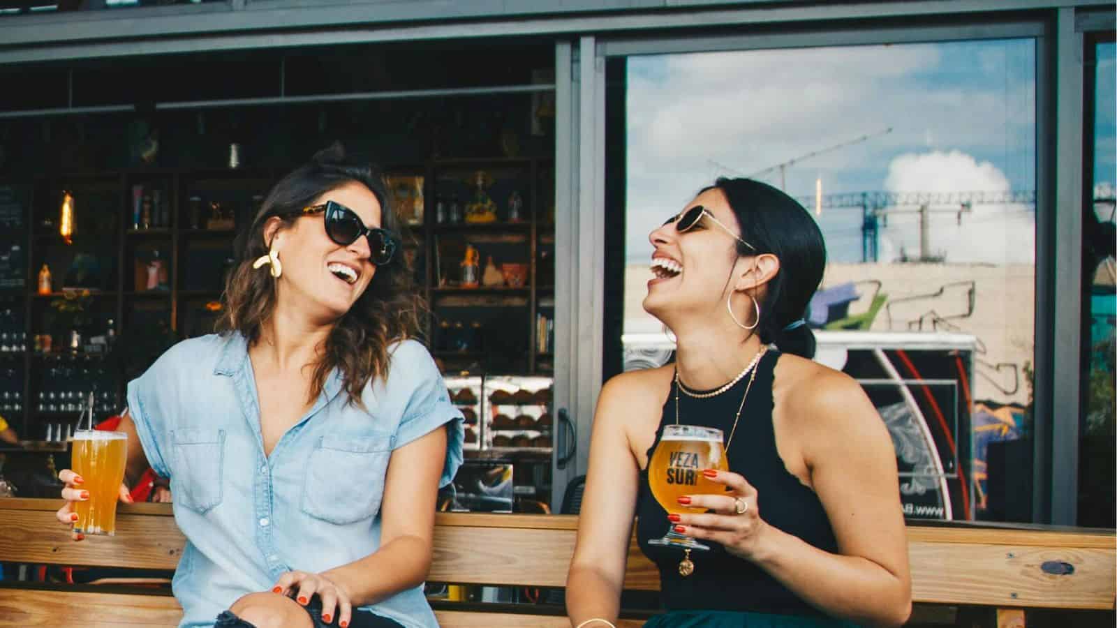 Two women are sitting outdoors on a wooden bench, laughing and holding drinks. Both women are wearing sunglasses; one is in a light blue shirt, and the other is in a black tank top. Shelves with bottles are visible in the background as they chat about their favorite alcohol rules.