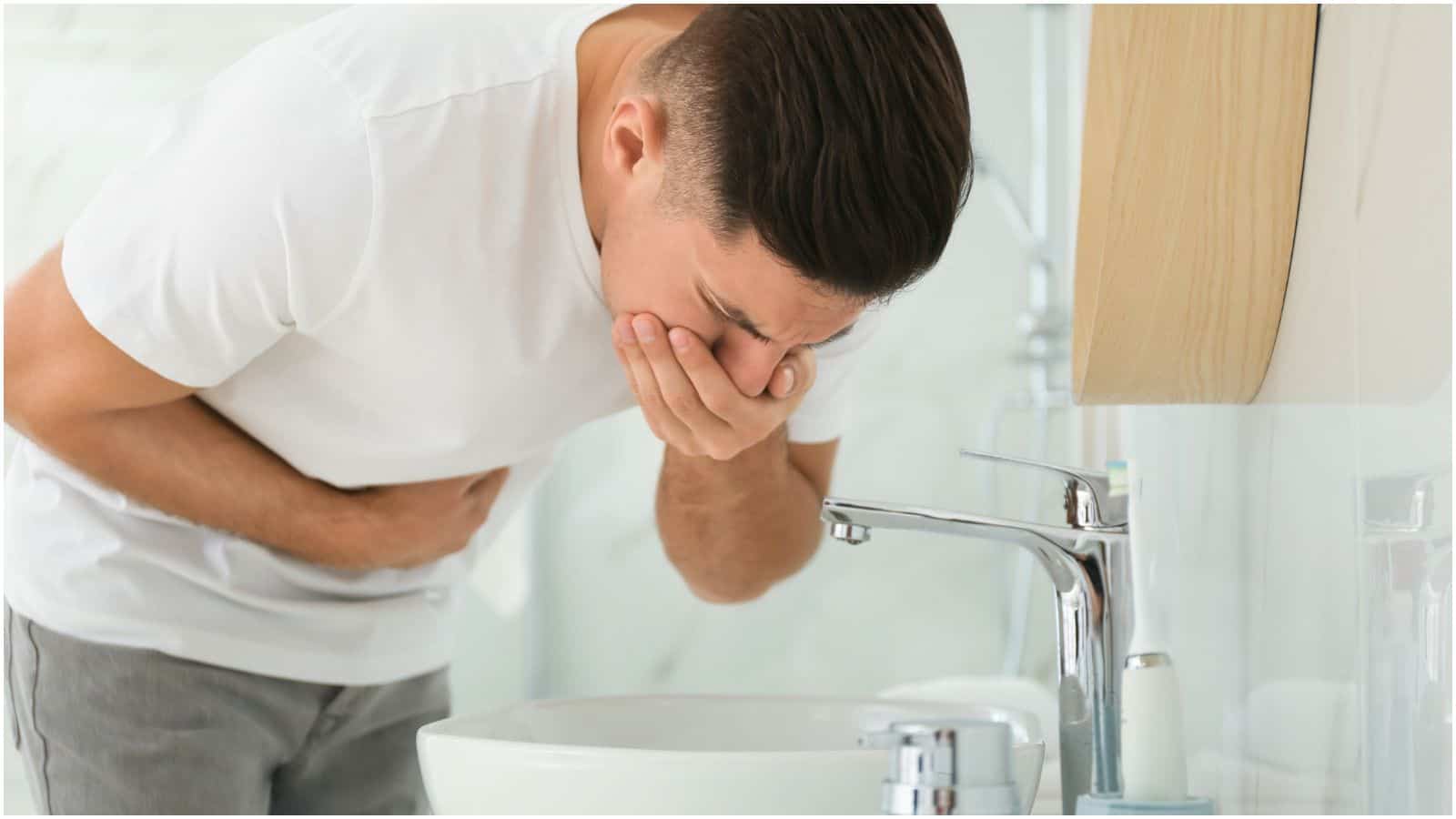A man in a white t-shirt stands in front of a bathroom sink, covering his mouth with one hand and holding his stomach with the other, appearing to feel unwell&mdash;perhaps a consequence of ignoring alcohol rules.