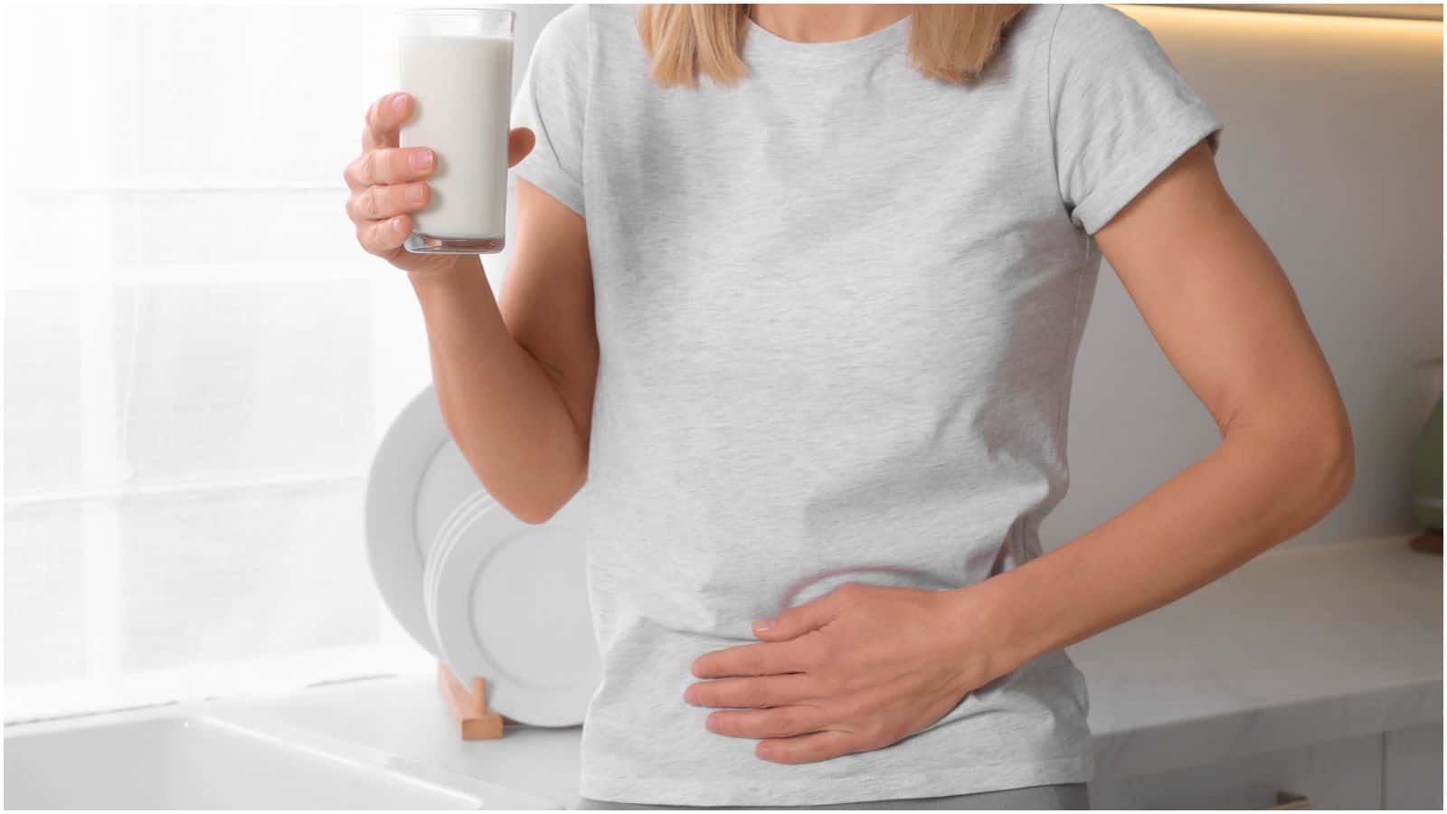 A person wearing a gray t-shirt is holding a glass of milk in their right hand while touching their abdomen with their left hand. They are standing in a kitchen next to a counter with white plates.