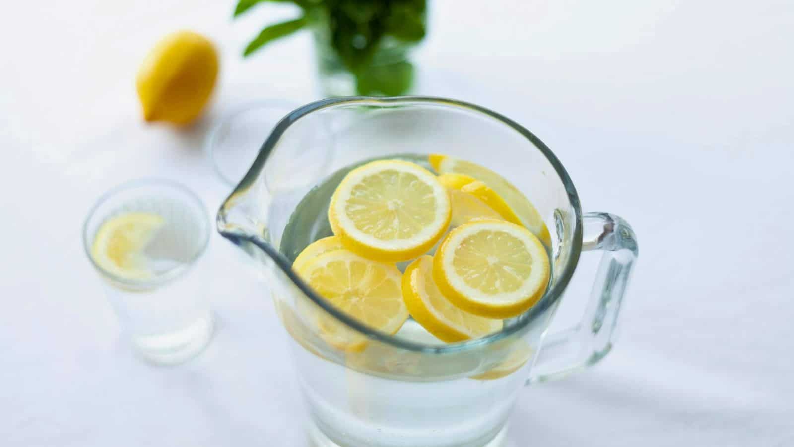 A glass pitcher filled with water and slices of lemon on a white surface. Beside it is a small glass cup with a lemon slice on the rim. In the background, there is a whole lemon and some green leaves.