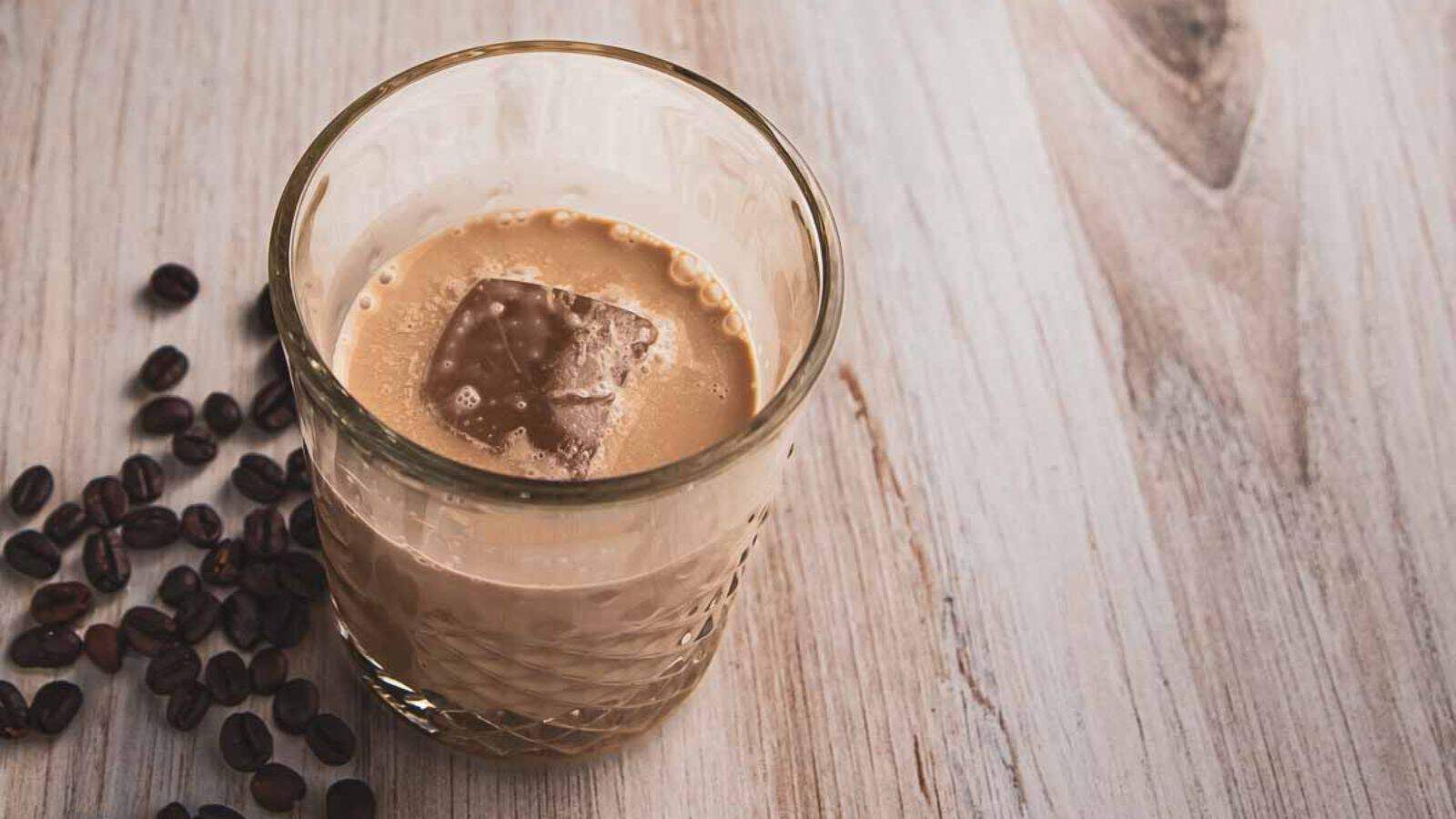 A glass with a cream-colored drink and a single ice cube sits on a wooden surface, hinting at the secret blend of alcohol within. Several coffee beans are scattered around the glass, adding an aromatic touch that breaks all the usual rules.
