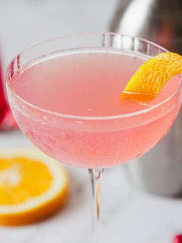 A pink cocktail garnished with a lemon twist in a coupe glass is the focal point. In the background are a cocktail shaker, a partially sliced orange, a small bottle of red liquid, and scattered cranberries on a marble surface.