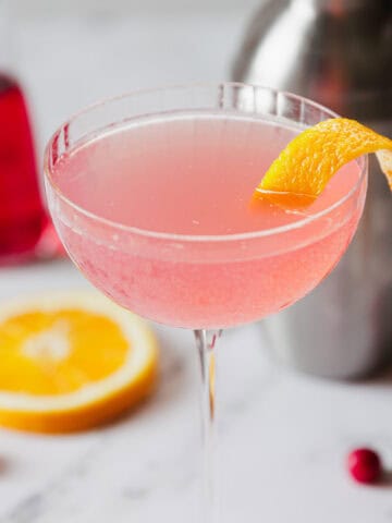 A pink cocktail garnished with a lemon twist in a coupe glass is displayed on a marble surface. In the background are a cocktail shaker, a slice of orange, fresh cranberries, and a small carafe with a red liquid.